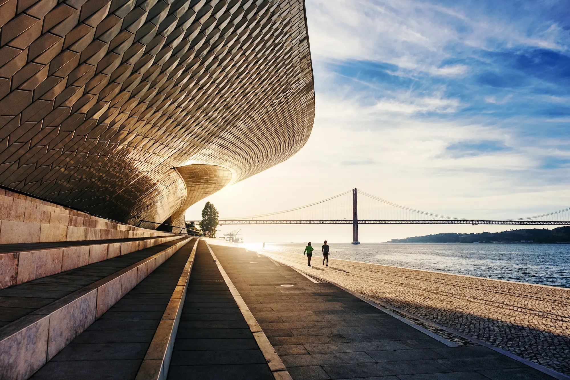 People walking along the Lisbon waterfront by the modern MAAT museum architecture with the 25 de Abril Bridge in the background, showcasing the city's active urban lifestyle.